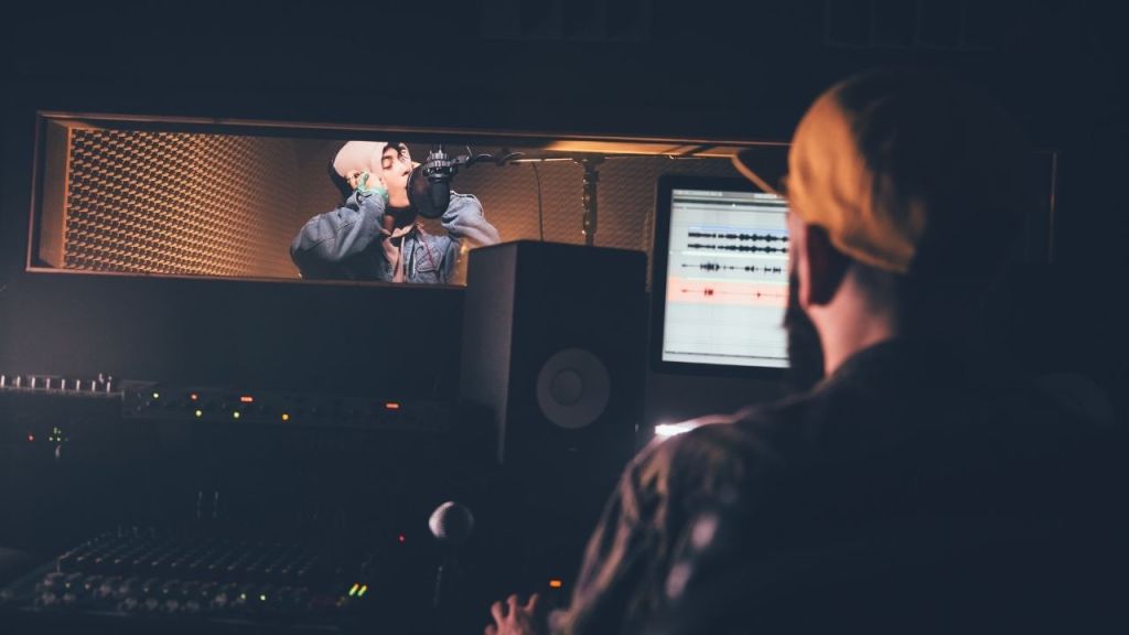 Music producer in a recording studio monitoring a vocalist through the control room window, illustrating how Auribus integrates seamlessly into existing workflows to audition toplines, test delivery, and build final takes efficiently.